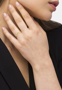 Goldfarbener Ring mit gewelltem Design, aus Metall gefertigt. Am Finger getragen, betont die glatte Textur und die reflektierende Oberfläche. Minimalistischer Stil.