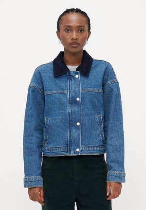Veste en denim présentant un délavage bleu, un col foncé contrastant, une fermeture à boutons et des poches latérales. Coutures visibles avec une coupe droite.