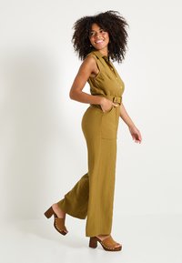 Une combinaison sans manches olive avec une ceinture à la taille, des poches et des jambes ample. Portée avec des talons ouverts marron ornés de clous.