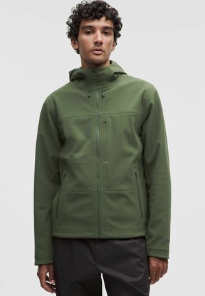 Uomo con giacca verde con cappuccio e zip e pantaloni neri, in piedi davanti a uno sfondo chiaro e uniforme.