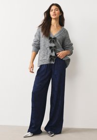 Cardigan gris en maille avec trois nœuds noirs, associé à un pantalon large marine à motif discret, et des chaussures argentées à bouts pointus.