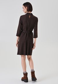 Robe marron longueur genou avec manches longues, cintrée à la taille. Présente un col montant, un dos avec ouverture en forme de goutte et des motifs subtils sur toute la surface.
