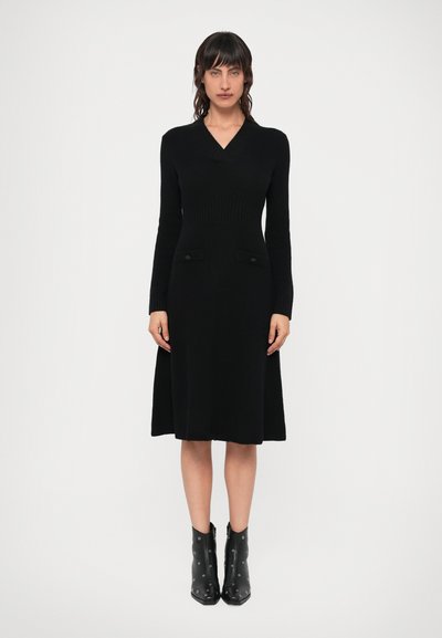 Schwarzes Strickkleid mit V-Ausschnitt, geripptem Oberteil und ausgestelltem Rock. Verfügt über zwei Vordertaschen mit Stoffakzenten. Getragen mit schwarzen Stiefeletten.