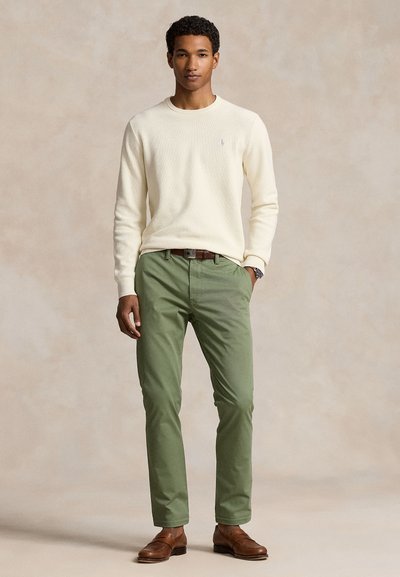 Kremowy sweter z dzianiny, zielone spodnie o dopasowanym kroju, brązowe skórzane buty i pasek. Prosty design, bez wzorów, z logo na swetrze.