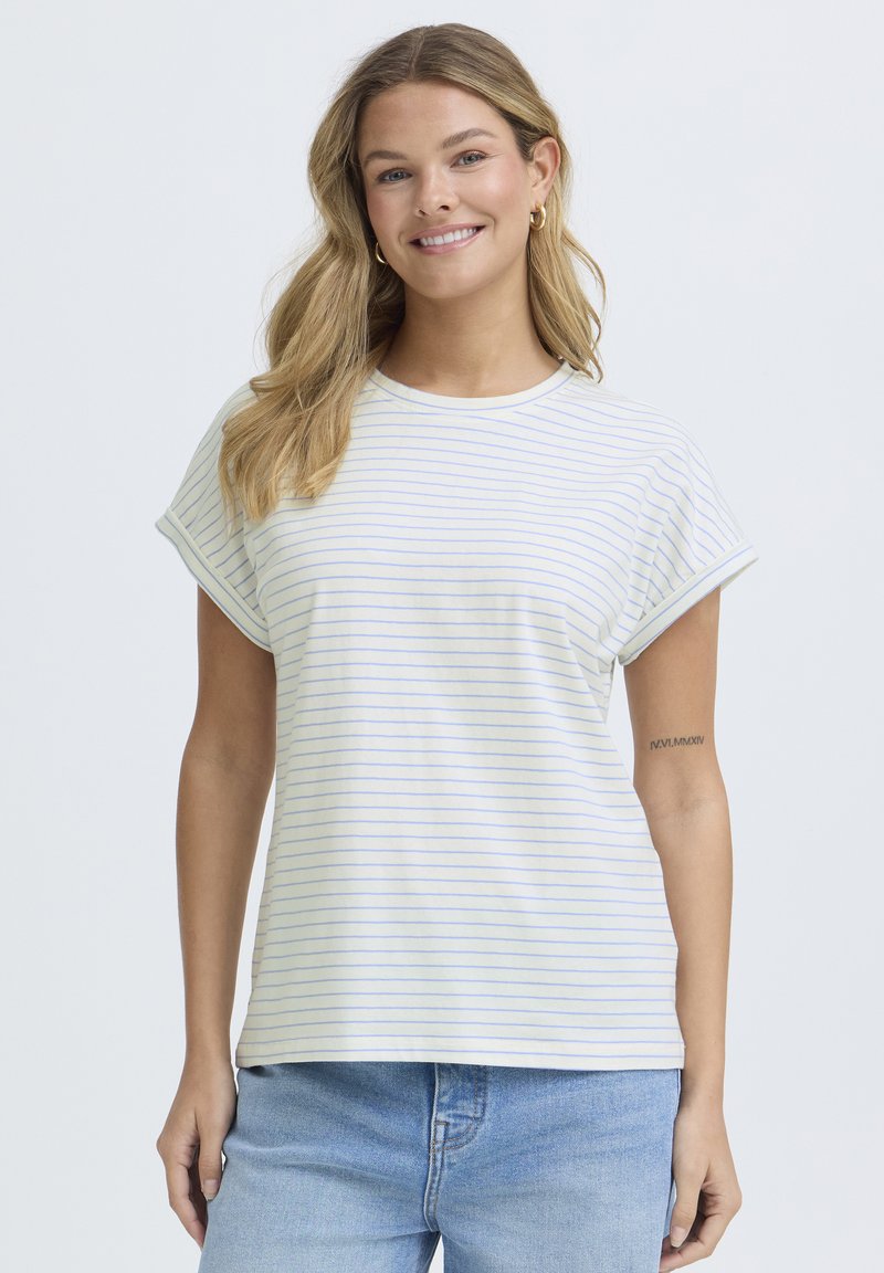 Femme souriante aux longs cheveux blonds portant des boucles d'oreilles dorées en forme d'anneau, une chemise légère à manches courtes rayée et un jean bleu, posant devant un fond uni.