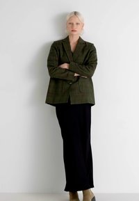 Blazer verde a quadri con un solo bottone, abbinato a una lunga gonna nera. Il modello ha i capelli corti e chiari e si trova davanti a uno sfondo bianco.