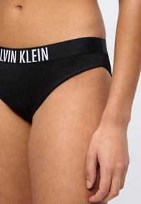 Femme portant des sous-vêtements noirs Calvin Klein avec le logo visible sur la ceinture, montrant le ventre, la hanche et la main droite sur fond blanc.
