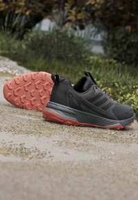 Chaussures de trail noires et grises avec semelles et lacets orange placées sur un chemin de gravier avec un feuillage vert en arrière-plan.