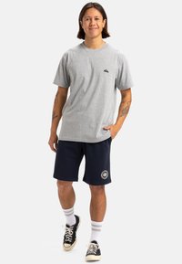 Camiseta de algodón gris con un pequeño logo negro en el pecho, combinada con pantalones cortos azul marino que presentan un parche con un logo redondo, calcetines blancos y zapatillas negras.