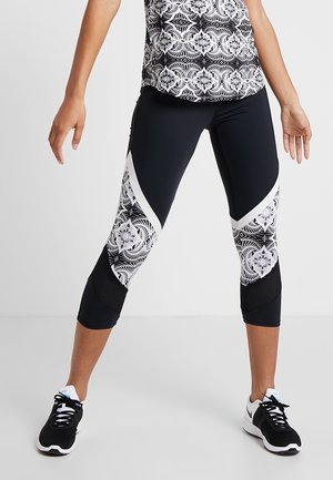 Frau trägt schwarz-weiß gemusterte, gekürzte Leggings und passendes Oberteil, steht mit gespreizten Beinen und entspannten Armen.