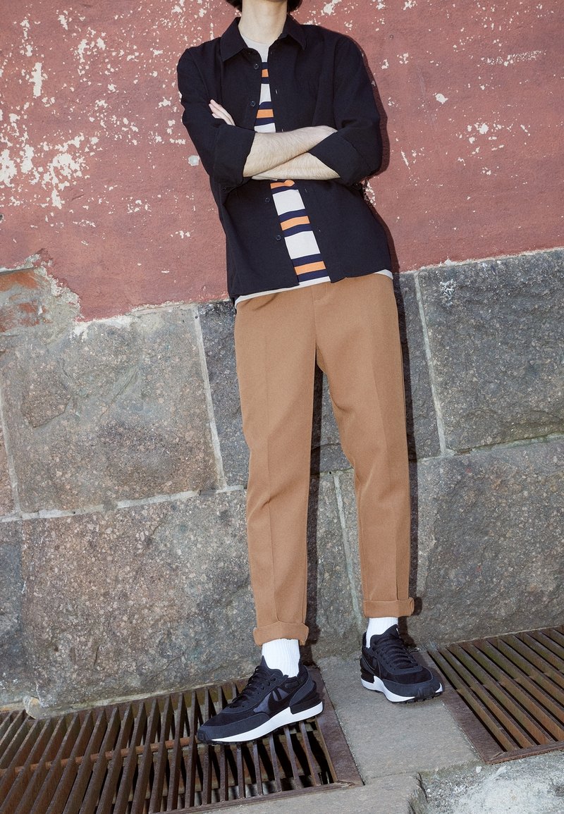 Black button-up shirt over a striped shirt, tan trousers, and black sneakers with white soles; against a textured red wall.