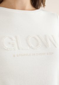 Vit bomull t-shirt med texturerad "GLOW" bokstäver i reflekterande pärlor och subtil beige text "& SPARKLE IN EVERY STEP" nedanför.