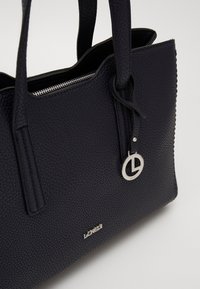 Sac en cuir texturé bleu foncé avec deux poignées, une fermeture zippée sur le dessus et un charm logo en argent accroché pour le détail.