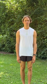 Jeune homme en débardeur blanc et short noir debout sur l’herbe avec des buissons verts feuillus en arrière-plan.