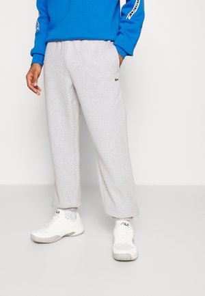 Homme portant un pantalon de survêtement gris clair, des baskets blanches FILA et un sweat bleu avec des bandes de logo blanches et noires sur les manches.
