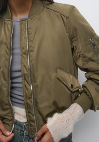 Persona vistiendo una chaqueta bomber verde oliva sobre una camiseta de rayas gris y jeans azules, con guantes de lana sin dedos en la mano izquierda.