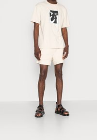 Homme debout portant un t-shirt graphique beige, un short beige assorti et des sandales noires à lanières sur un fond uni.