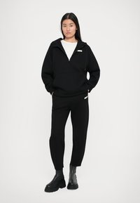 Mladá žena stojí s rukami v vreckách, oblečená v čiernej zip-up mikine, zodpovedajúcich nohaviciach, bielej košeli a čiernych robustných čižmách na bielom pozadí.