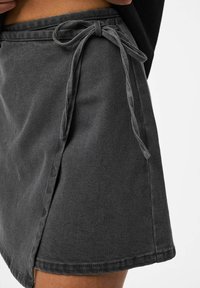 Gros plan sur une jupe en denim gris foncé avec fente avant et ceinture nouée, portée par une personne à la peau brune et un haut noir.