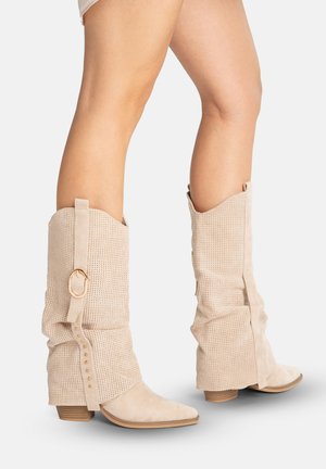 Beine mit beigen, gelochten, lässigen kniehohen Stiefeln mit niedrigen Holzabsätzen und goldfarbenem Schnallenriemen-Detail vor weißem Hintergrund.