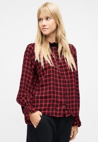 Ξανθιά γυναίκα που φοράει μια κόκκινη και μαύρη καρό μπλούζα με μακριά μανίκια και βολάν στα μανίκια, καθώς και μαύρο παντελόνι, κοιτάζει προς τα αριστερά της.