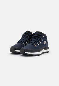 Zapatos de senderismo de media altura en malla azul marino con acentos negros, ojales plateados y suela blanca. Presentan materiales duraderos y una parte superior texturizada.