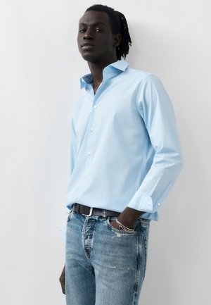 Uomo dalla pelle scura con dreadlocks che indossa una camicia azzurra con bottoni e jeans blu, con una mano in tasca su sfondo bianco.