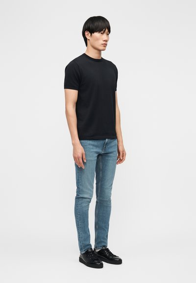 Νεαρός άνδρας όρθιος, φοράει απλό μαύρο κοντομάνικο μπλουζάκι, ανοιχτόχρωμο τζιν στενής γραμμής και μαύρα αθλητικά παπούτσια, με φόντο λευκό χρώμα.