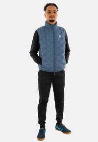 Gilet imbottito blu con design a trapunta verticale, collo alto e chiusura con zip, abbinato a pantaloni neri e sneakers navy con accenti blu.