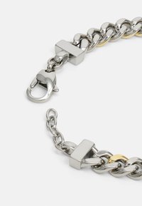 Pulseira de corrente metálica com elos alternados em prata e ouro, um fecho retangular e um fecho de lagosta com um acabamento brilhante.