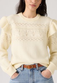 Pull en maille crème avec des manches bouffantes, présentant un motif en dentelle décoratif sur la poitrine, associé à un jean bleu taille haute et une ceinture marron.