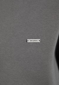 T-shirt grigio realizzato in un tessuto liscio simile al cotone, con un piccolo marchio metallico argentato con la scritta "SMILODOX" attaccato davanti.