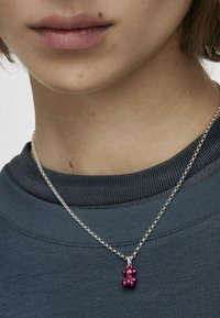 Collar de cadena de plata con un colgante de oso de goma morado, sobre un escote de sudadera de textura azul oscuro.