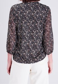 Blouse noire à motifs floraux composée d'un tissu transparent, avec des manches 3/4, un col rond et un ourlet froncé. Le design présente de petits motifs floraux roses et blancs.