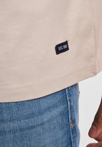 T-shirt in cotone beige con etichetta nera con "EST. 1990" vicino all'orlo, abbinato a jeans in denim blu. Cuciture visibili all'orlo.