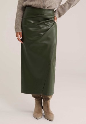 Donna che indossa una gonna midi in pelle verde scuro a vita alta abbinata a stivali in camoscio beige e a un maglione beige lavorato a maglia.
