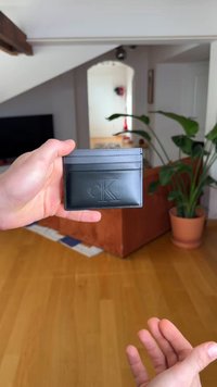 Zwarte leren kaarthouder met gegraveerd logo, vier vakken en een slank rechthoekig ontwerp. Getoond tegen een eenvoudige binnenachtergrond.