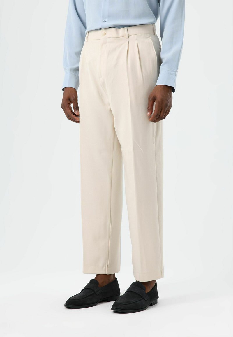Beige pleated trousers made from a lightweight fabric, featuring a straight cut and subtle creases. Paired with dark slip-on shoes.
