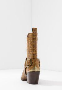 Botín de tobillo de piel sintética marrón con detalle de hebilla lateral, puntera afilada y tacón apilado. Presenta patrones texturizados y una suela lisa.