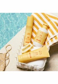 Wella Invigo Sun Care -tuotteet keltaisissa putkissa ovat valkoisessa korissa. Mukana on suihke, shampoo ja hoitoaine, valkoisella pyyhkeellä, jossa on raidallisia yksityiskohtia.