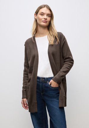 Brun strikket cardigan med åbent front, ribbede manchetter og sidelommer, båret over en hvid top og parret med blå jeans.