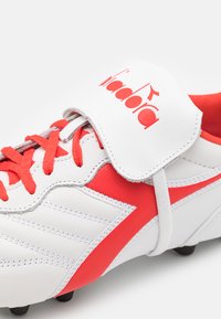 Scarpa da calcio bianca con accenti rossi, caratterizzata da pelle testurizzata, colletto imbottito e lacci a contrasto. Logo visibile sulla linguetta.
