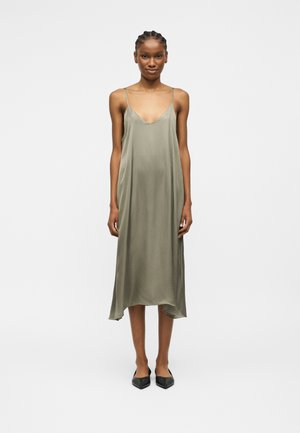 Pikk naine punutud juustega, kandes õlgu paljastavat, lõdva istuvusega oliivs-rohelise tooni midi-kleiti ja musti terava ninaosaga kingi, seisab tavalise tausta ees.