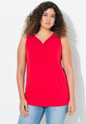 Top sans manches rouge avec un décolleté en V, présentant des détails froncés au centre. Tissu doux, coupe décontractée, associé à un jean bleu clair.
