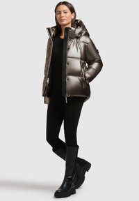 Skinner metallisk pufferjakke i mørkegrå med høj krave, frontknapper og sidelommer, parret med sorte tætsiddende bukser og støvler til knæene.