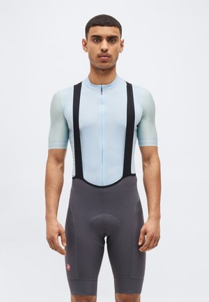 Mann trägt hellblaues kurzärmeliges Radtrikot und schwarze, gepolsterte Trägerhose, steht vor weißem Hintergrund und schaut nach vorne.