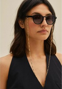 Černé kulaté sluneční brýle s metalickou zlatou řetízkou. Brýle mají tmavé čočky a štíhlé, rovné nohy. Řetízek má prodloužené články pro podporu.
