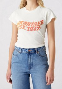 Bílá bavlněná tričko s oranžovým grafickým potiskem s nápisem "Wrangler Est. 1947." Krátké rukávy a přiléhavý střih, spárované s modrými džíny.