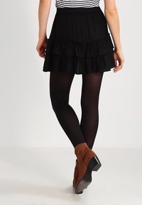 Femme portant une mini-jupe noire à volants, des collants opaques noirs et des bottes à chevilles en daim marron, vue de dos.