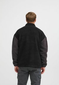 Chaqueta de felpa negra y gris con un acabado texturizado y esponjoso, que presenta un cuello alto y mangas de contraste. Ajuste casual con mangas largas.
